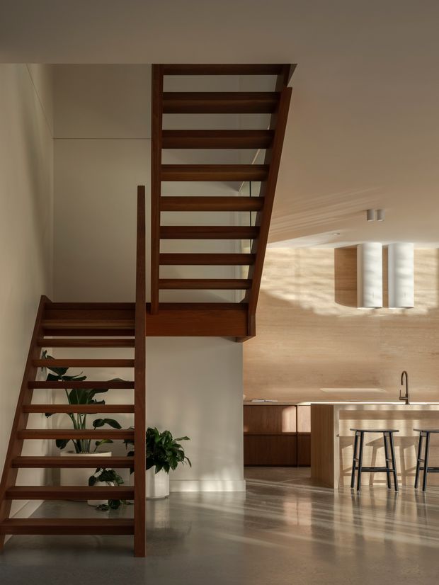 A warm, contemporary open-plan interior featuring a sculptural open-riser timber staircase in the foreground, leading toward a kitchen with dark wood cabinetry, a pale stone island, bar stools, and a brushed metal tap. Sunlight creates strong shadows across the timber-lined kitchen wall and polished floor, while indoor plants add greenery beneath the stairs.