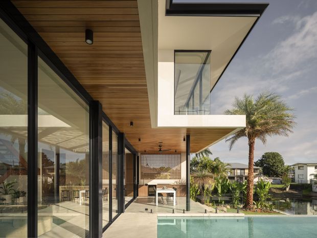Contemporary multi-level residence with extensive black-framed glazing, warm timber soffits, white rendered volumes, and a covered outdoor entertaining area overlooking a swimming pool and landscaped garden. The image emphasizes the rear facade, covered terrace, and strong indoor-outdoor connection.
