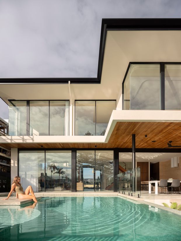 A contemporary multi-level residence viewed from a pool courtyard. The home features extensive floor-to-ceiling glazing with dark metal frames, projecting roof planes, warm timber soffits, an open-plan entertaining area with dining furniture, and a turquoise swimming pool in the foreground.