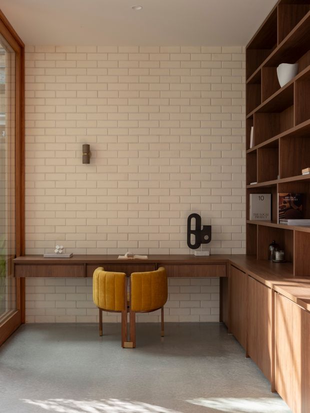 A refined home office with a built-in dark timber desk and extensive shelving, set against a pale cream brick feature wall. Two mustard upholstered chairs sit at the desk, with warm timber-framed glazing on the left and soft natural light across a smooth grey floor.