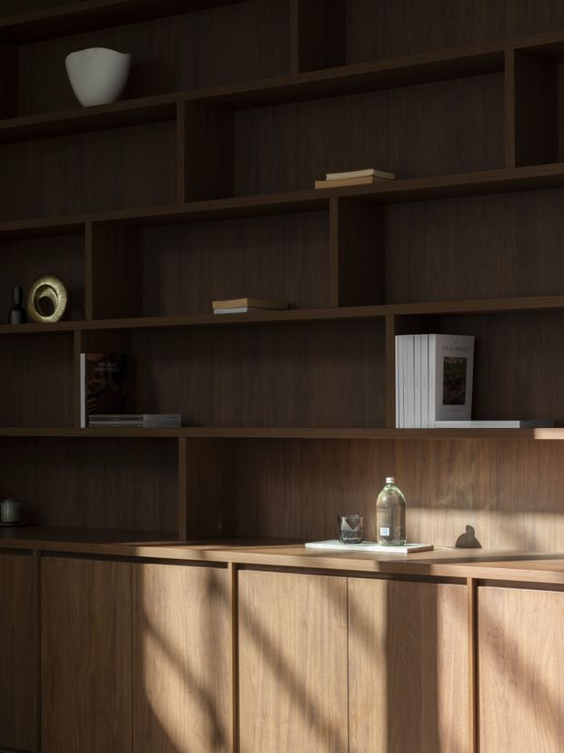 A dark timber built-in shelving and cabinetry wall fills the frame, with asymmetrical open shelves displaying a few books and understated decorative objects. Lower closed cupboards form a continuous sideboard, illuminated by warm directional sunlight and strong shadows. The composition is refined, minimal, and strongly wood-focused.