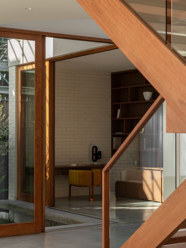 A warm, light-filled contemporary interior viewed through an open timber-framed glazed doorway. The space features a pale painted brick wall, dark built-in shelving, a compact desk with mustard upholstered chairs, polished concrete flooring, and a prominent timber stair structure with glass balustrade in the foreground. An outdoor courtyard and additional glazing are visible beyond.