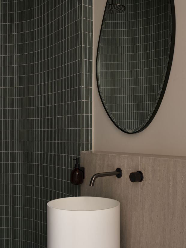 Close-up of a refined contemporary bathroom vanity area. A white cylindrical vessel basin sits against a pale stone-look wall panel, with a dark wall-mounted tap and matching control. The adjacent shower wall is clad in dark green vertically gridded tiles, and a large organic oval mirror with a black frame reflects the tiled shower area and overhead shower fitting. The palette is dark green, warm beige, white, and black, with a calm, moody, luxurious feel.