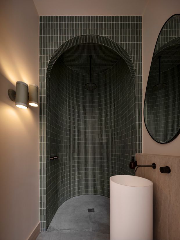 A moody contemporary bathroom featuring a tall arched walk-in shower lined with dark green narrow-format tiles, a curved tiled ceiling, grey concrete-look shower floor, wall-mounted black fixtures, a sculptural white cylindrical basin, and a large rounded mirror. Warm wall lighting creates a refined, atmospheric feel.