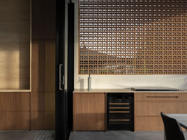A refined contemporary kitchen or dining area featuring dark timber cabinetry, a pale stone benchtop, integrated wine storage, a black-framed glazed door, and a large decorative woven timber screen. The space has a warm, textural material palette with dramatic sunlight and shadow.