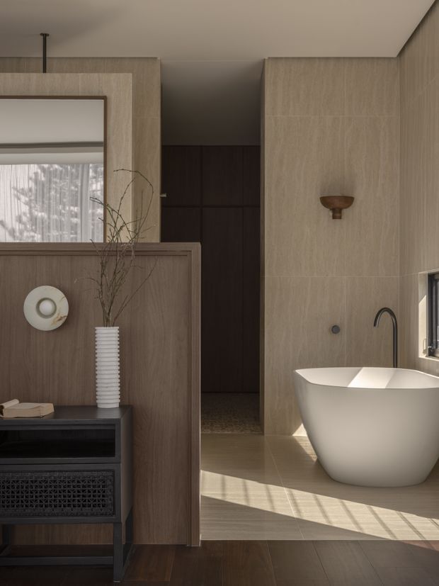 A warm, contemporary bathroom with a freestanding white bathtub, matte black wall-mounted tapware, and a small wall sconce. Large light beige stone-look wall and floor surfaces contrast with dark timber cabinetry and a partial timber privacy screen. Natural light enters through a window, creating strong diagonal shadows across the floor. A dark doorway or passage is visible behind the bath, while a console and decorative vase occupy the foreground.