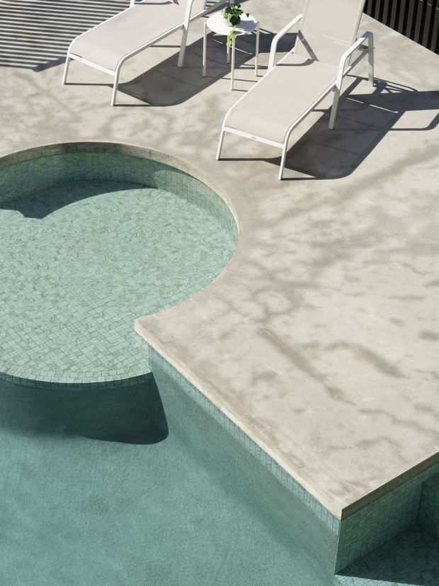 A sunlit pool terrace with sculptural curved pool edges, pale green mosaic tile lining, and a light concrete deck. Two minimalist white lounge chairs and a small round side table sit beside the pool, with strong architectural shadows creating a clean, contemporary outdoor atmosphere.