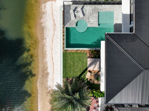 Aerial view of a contemporary waterfront property beside a sandy shoreline. The image prominently shows a turquoise swimming pool with a circular spa, paved pool terrace, manicured lawn, tropical planting, and a dark metal hipped roof.
