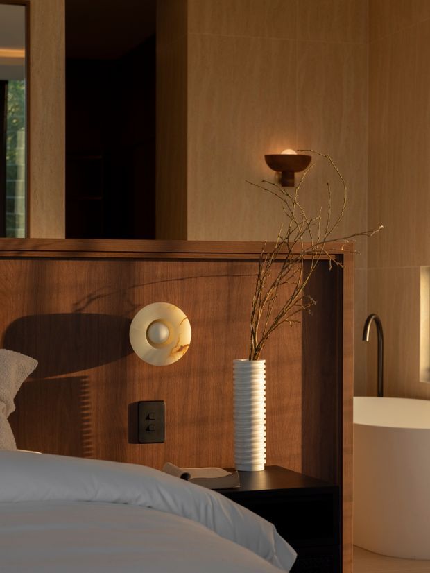 Warmly lit bedroom vignette featuring a timber-panelled headboard and wall, bedside table, sculptural wall light, decorative vase with branches, and a partially visible freestanding bathtub in an adjoining bathroom area. A dark doorway and mirror or glazing appear to the left.