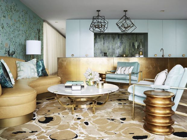 A richly styled open-plan living room with a compact kitchen at the rear. The space features a curved tan sectional sofa, pale blue lounge chairs, a round white coffee table with a brass base, a sculptural brass side table, a patterned area rug, floral wallpaper, white cabinetry, a metallic gold-toned kitchen island or partition, and a dark textured splashback. Geometric black pendant lights hang above the seating and kitchen area.