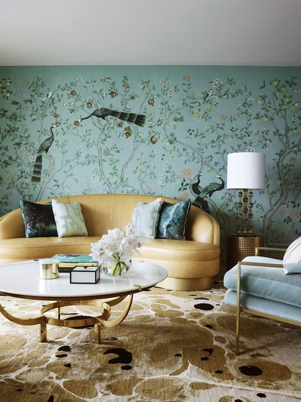 A richly layered living room with a curved mustard-gold sofa, pale blue armchair, sculptural oval coffee table, patterned area rug, floral-and-bird wallpaper, decorative cushions, and a table lamp. The room combines botanical motifs, saturated jewel tones, metallic accents, and rounded mid-century-inspired furniture in an elegant, playful composition.