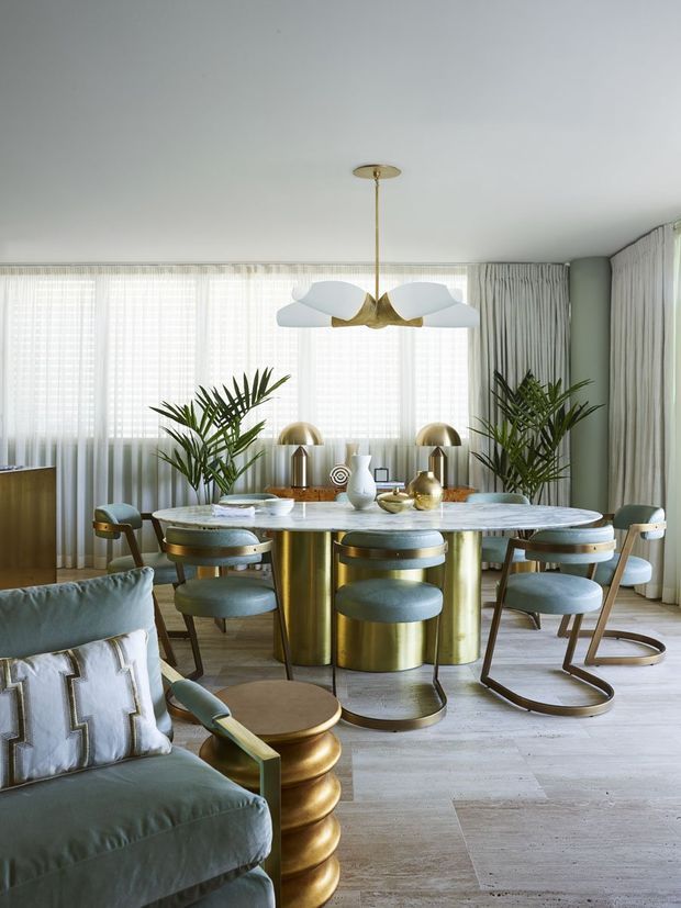 A refined dining and living area with a large round marble dining table, sculptural brass pedestal base, upholstered blue-grey dining chairs, and a statement white-and-brass pendant light. Floor-to-ceiling sheer curtains surround the room, with tall indoor palms, brass table lamps, and a green upholstered sofa in the foreground. The space has a polished, luxurious mid-century-inspired appearance with soft natural light and warm metallic accents.