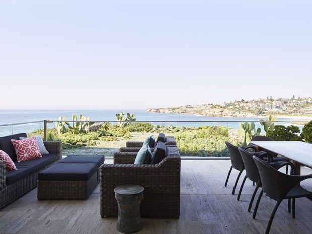 A spacious coastal outdoor terrace with dark woven lounge seating, a dining table and chairs, and a glass balustrade overlooking the ocean and a sandy shoreline. The terrace has weathered grey timber decking, low coastal planting, and a bright open atmosphere.