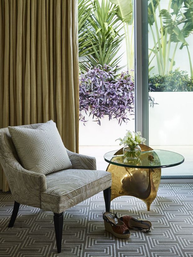 A refined sitting area beside full-height glazing, featuring a patterned upholstered armchair, geometric grey rug, pleated floor-to-ceiling curtains, and a round glass-topped side table with an ornate gold sculptural base. Lush tropical planting is visible outside through the glazing. The interior has a polished, luxurious, pattern-rich character with strong geometric motifs and warm metallic detailing.