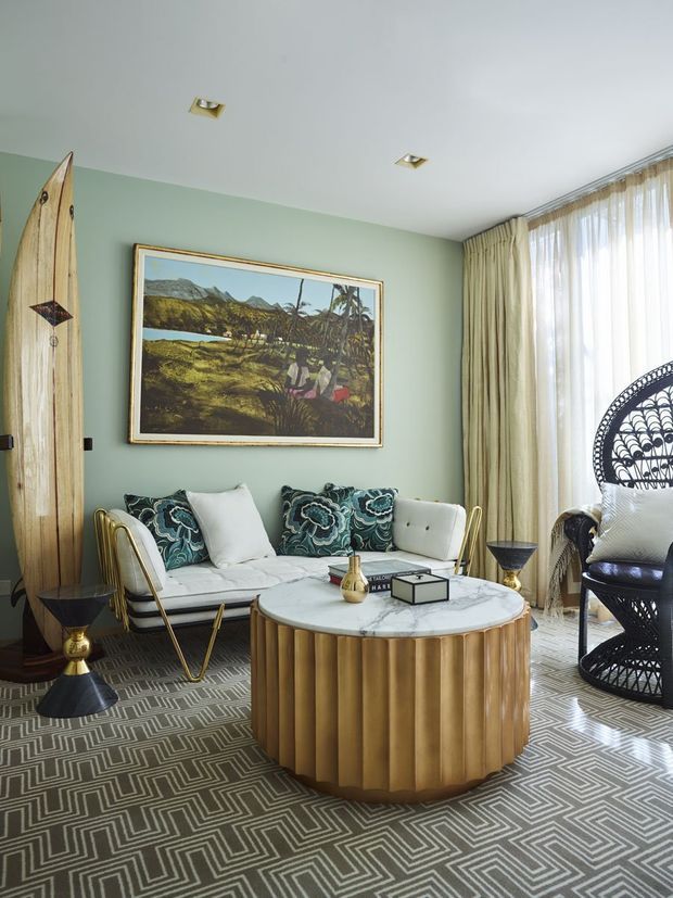 A stylish living room with sage-green walls, sheer floor-to-ceiling curtains, a white sofa with patterned teal cushions, and a prominent round coffee table with a veined white stone top and fluted timber base. A large framed tropical landscape artwork, sculptural black-and-gold side tables, a decorative surfboard, a wicker peacock chair, and a geometric patterned rug create a bold, eclectic interior.