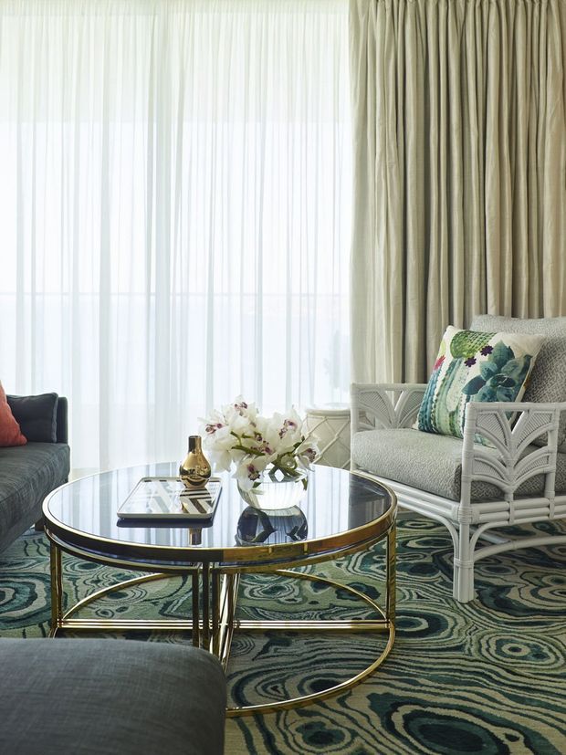 Elegant living room with a large round glass coffee table on a gold-toned metal frame, upholstered seating, a white wicker-style armchair with a botanical cushion, full-height sheer and taupe drapery, and a bold dark green patterned carpet. The room is softly lit and richly layered, combining classic furnishings with glamorous metallic detailing and botanical accents.