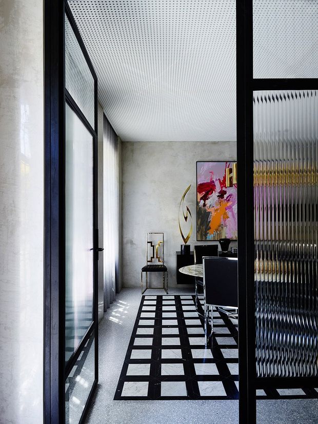 A refined dining room viewed through black-framed glazed doors. The space features pale textured walls, a patterned white ceiling, a monochrome geometric floor treatment, a round dining table with dark upholstered chairs, sculptural decor, and a colourful abstract artwork. Ribbed and translucent glazing adds texture and filtered light.