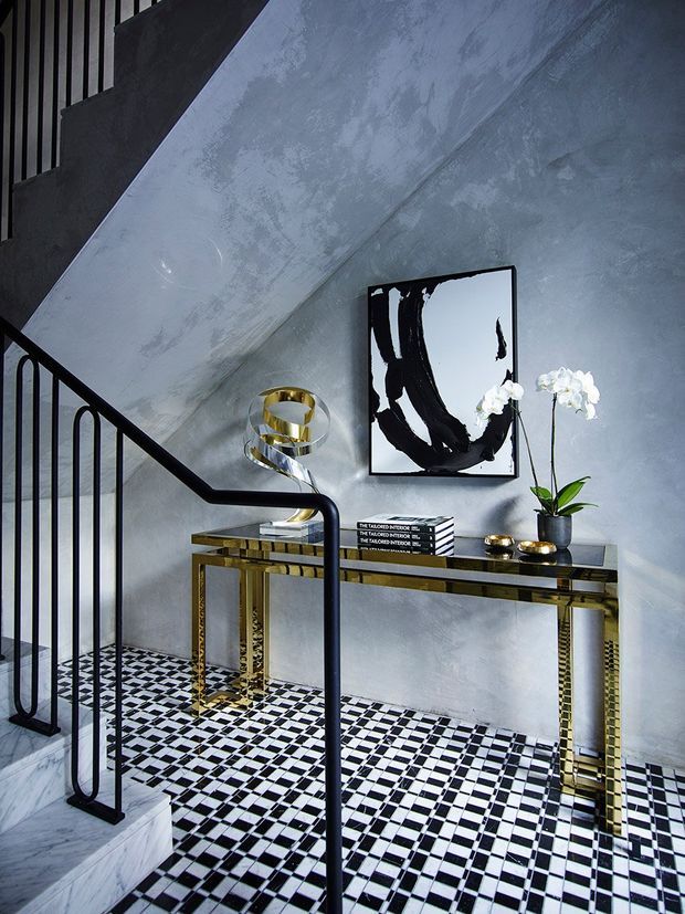 A dramatic stairway landing with a black-and-white checkerboard tile floor, pale textured plaster walls, black metal balustrades, and a slim console table with reflective gold-toned legs. The console is styled with a sculptural chrome-and-gold lamp, stacked books, small gold objects, a potted white orchid, and bold black-and-white abstract artwork. The space has strong geometric contrast and an elegant, glamorous feel.