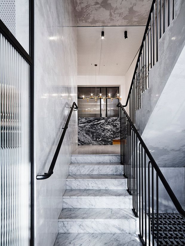 A refined contemporary stairway with full-height white and grey marble-clad walls and stair treads, black metal balustrades and handrails, and a perforated ceiling. At the landing, a dramatic black-and-white veined stone feature wall and linear pendant lighting create a bold focal point.