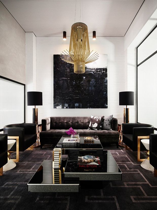 Sophisticated living room with a dark velvet sofa, matching black armchairs, mirrored black coffee tables, black-and-grey geometric carpet, and tall black floor lamps with brass-toned stems. White painted brick walls and a perforated acoustic ceiling provide contrast, while a large black abstract artwork and sculptural brass pendant light form dramatic focal points. Full-height glazed partitions with dark frames flank the room.