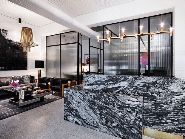 A refined contemporary commercial reception or hospitality interior with a dramatic black-and-white veined stone reception counter in the foreground. Behind it are black-framed glass and ribbed-glass partitions, a lounge seating area with dark furniture, a large monochrome artwork, sculptural pendant lighting, and an exposed white mechanical duct beneath a perforated ceiling.