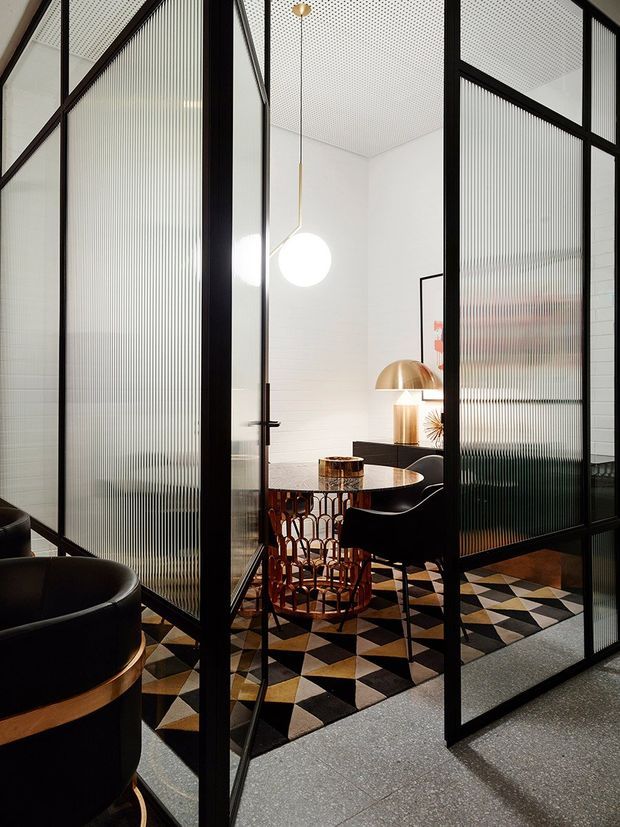 A refined interior shown through black-framed ribbed glass partitions and a partially open glass door. The space contains a small round dining or meeting table, sculptural black chairs, a brass-toned dome lamp, a spherical pendant, white tiled walls, and a bold geometric black, cream, and ochre rug over a grey floor. The composition emphasizes screened zones, graphic pattern, and warm metallic accents.