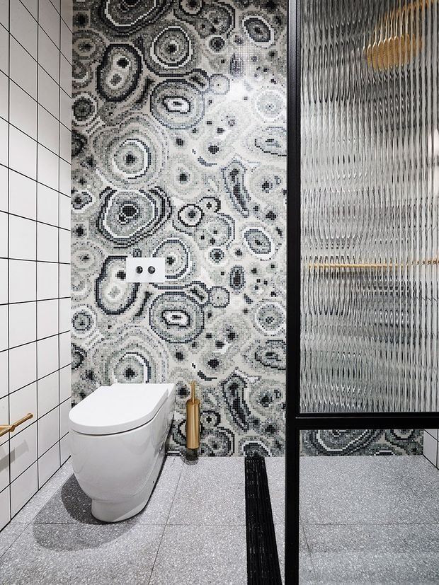 Compact contemporary bathroom featuring a wall-hung toilet, white square-grid wall tiles, a dramatic black-and-white abstract patterned feature wall, grey speckled floor tiles, and a black-framed ribbed glass partition or door. Brass fixtures add a warm accent.