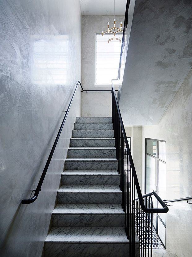 A bright, narrow interior stairway with grey veined marble-clad treads and risers, white textured plaster walls, and slender black metal handrails and balustrades. Tall windows bring in abundant natural light, while a small brass-and-white chandelier hangs above the stairwell. The stair continues down to a lower level visible at right.