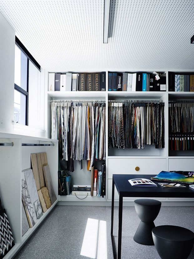 Bright, compact materials and textile sample library with floor-to-ceiling built-in shelving, hanging fabric swatches, books, material boards, and a dark worktable with stools. The space has white cabinetry, a perforated acoustic ceiling, large black-framed windows, and a cool grey floor.