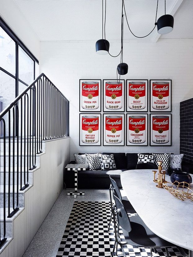 A striking open-plan dining and living area with a bold black-and-white palette. The space features a long white marble dining table with black chairs, a black sofa with graphic patterned cushions, checkerboard rugs and side tables, framed Campbell's Soup artwork, black globe pendant lights, a black metal stair balustrade, and large industrial-style windows. White painted brick walls and a perforated white ceiling create a bright, graphic interior.