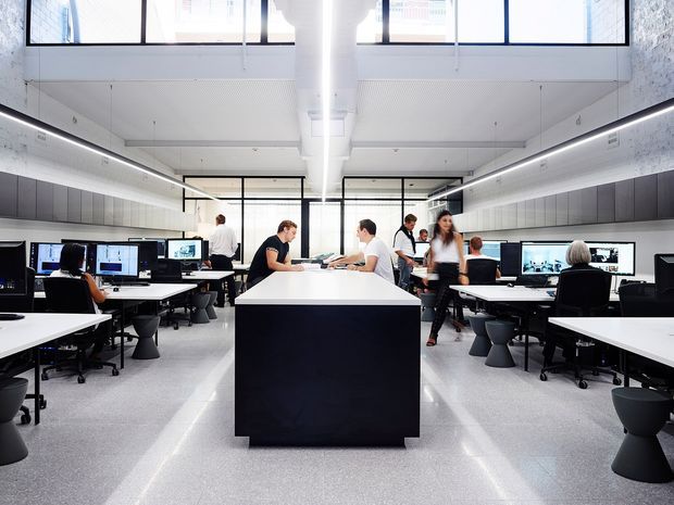 Bright contemporary open-plan commercial workspace with rows of computer workstations arranged around a large central island table. The room has a high ceiling, clerestory windows, extensive glazing, linear integrated lighting, pale speckled flooring, dark built-in cabinetry, and multiple people working at desks.