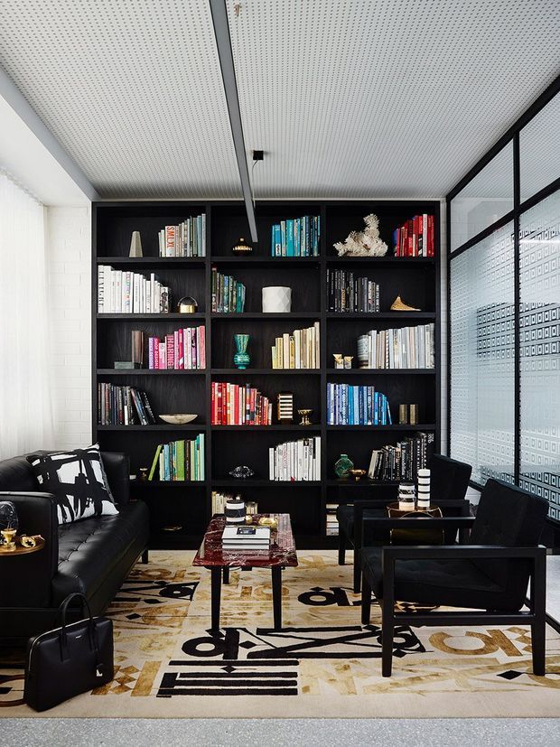 A sophisticated library-style living room with a floor-to-ceiling black built-in bookcase filled with colourful books and decorative objects. The room features black leather seating, a low glass-topped coffee table, a bold graphic patterned rug, and black-framed glazed partitions with a geometric privacy pattern. A perforated acoustic-style ceiling and white brick walls provide contrast to the dark joinery and furniture.