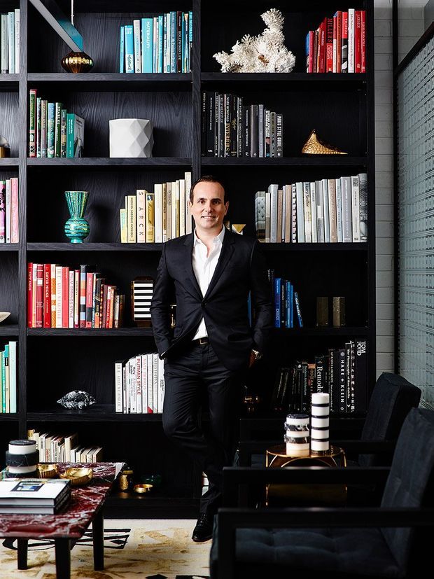 A dark, sophisticated living room or library-style lounge dominated by floor-to-ceiling black built-in bookshelves filled with colourful books and decorative objects. A man in a black suit stands in front of the shelving. The room includes black lounge seating, a low coffee table with books and decorative items, a patterned rug, and a textured grey block wall on the right. The styling is dramatic, refined, and highly curated.