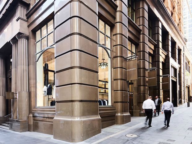 Street-level view of an imposing urban commercial facade with large rusticated stone-clad piers, classical fluted columns, deep horizontal banding, tall glazed shopfront windows, and ornate suspended signage. Pedestrians walk along the wide paved sidewalk, while modern high-rise buildings are visible in the background.