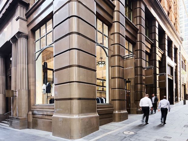 Street-level view of a refined commercial retail facade on a city sidewalk. The building features large fluted classical columns, substantial rounded pilasters, dark metal detailing, large display windows, stone or rendered masonry cladding in warm beige-grey tones, and visible retail interiors. Pedestrians pass along the paved urban frontage.