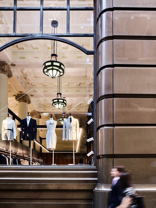 Street-level view into a refined fashion retail interior through a large glazed storefront. The shop features displayed mannequins, clothing racks, dark timber or metal joinery, ornate decorative ceiling detailing, large hanging pendant lights, and a substantial fluted stone or masonry facade column in the foreground. The architecture combines historic detailing with a polished contemporary retail fit-out.
