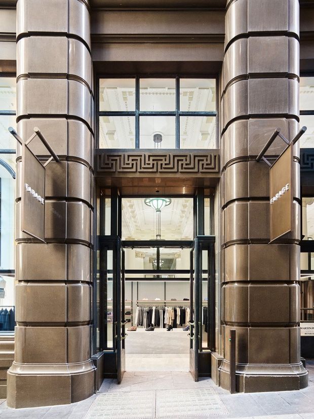 Symmetrical view of an ornate commercial retail entrance framed by large brushed-metal clad columns with horizontal segmented detailing. Dark metal and glass double doors open into a bright clothing boutique, with a Greek key frieze above the entry, decorative classical ceiling details, and patterned stone paving at the threshold.