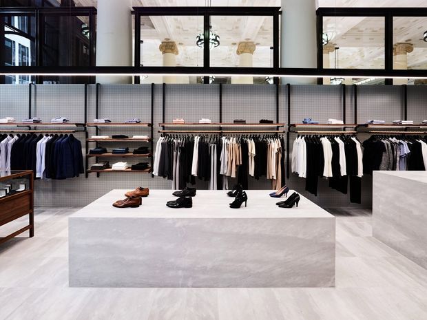 A refined contemporary clothing retail store interior with clothing racks arranged against grey perforated wall panels and a central pale stone display plinth showcasing shoes. The space features pale stone-look flooring and display surfaces, dark timber or metal-framed glazed partitions, white structural columns, and a bright, minimal monochrome palette. Reflections through the upper glazing reveal ornate ceiling detailing and columns in an adjacent or reflected space.