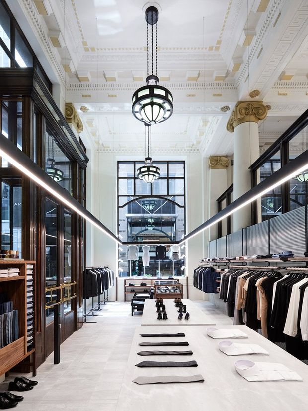 A refined high-end clothing retail interior with a long central display table, folded garments and shoes, clothing racks along both sides, dark timber and glass cabinetry, and a dramatic double-height atrium. The space features ornate white classical detailing, coffered and decorative ceiling mouldings, large fluted columns with gilded Corinthian capitals, black-framed glazing, and contemporary ring pendant lights. The palette is predominantly white, cream, black, and dark timber, combining heritage architectural character with a polished modern retail fit-out.