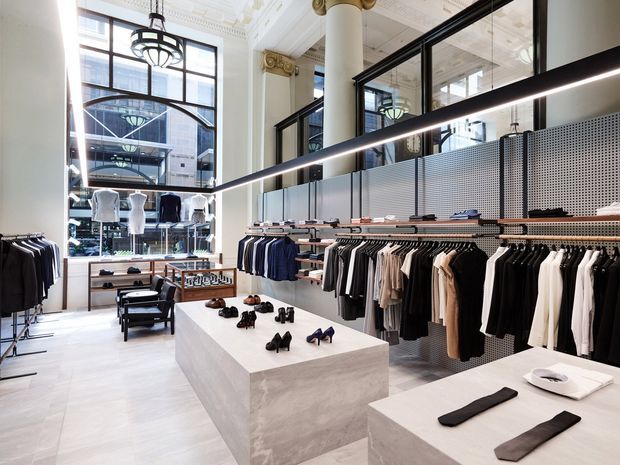 Bright high-end fashion retail interior in a heritage-style commercial space, with clothing racks, shoe and accessory display plinths, perforated metal wall panels, large black-framed glazing, ornate decorative ceiling detailing, classical columns, and contemporary linear lighting.
