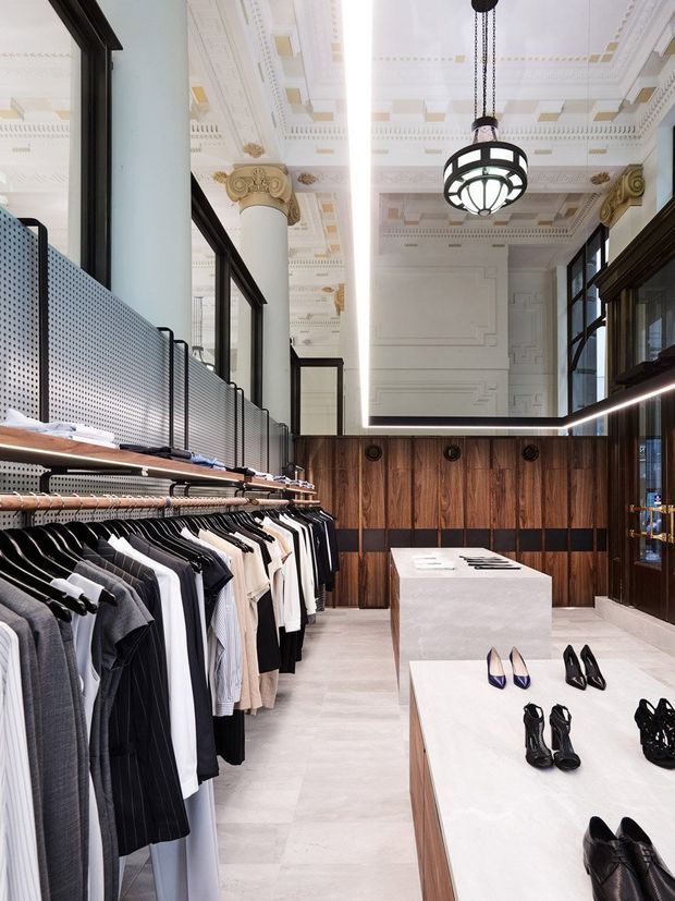 A refined clothing and footwear retail interior combining heritage architectural detailing with contemporary merchandising. The space features ornate white coffered ceilings, classical columns with gold-accented capitals, dark-framed glazed partitions, warm vertical timber wall panelling, black trim, pale stone flooring, and minimalist white display plinths. Clothing racks line the left side while shoes are arranged on central tables.