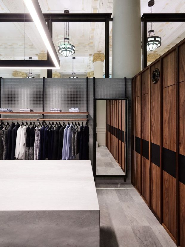 Interior view of a refined clothing retail space, featuring dark timber wall cabinetry with black horizontal insets, perforated metal clothing displays, a large pale stone or concrete counter, grey tiled flooring, and a full-height mirror. Decorative historic architectural elements, columns, ornate ceiling details, and black-and-white pendant lights are visible above the contemporary fit-out.