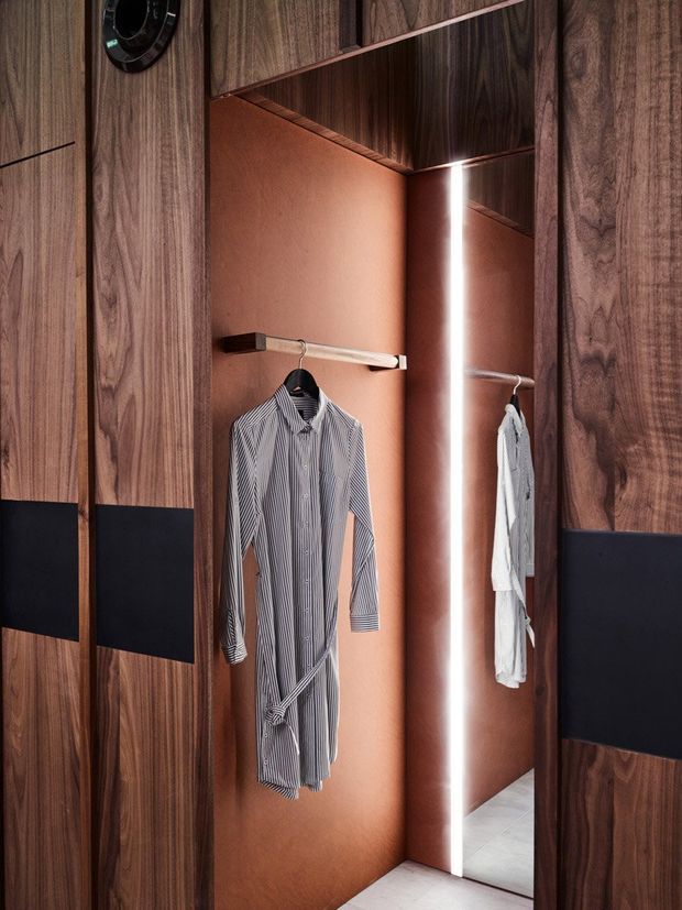 A close-up view of a refined walk-in wardrobe with dark walnut-toned timber cabinetry and black inset panels. A warm terracotta-toned wardrobe interior features hanging rails with shirts, a full-height mirror, and integrated vertical LED lighting. The space has a dark, luxurious, highly tailored appearance.