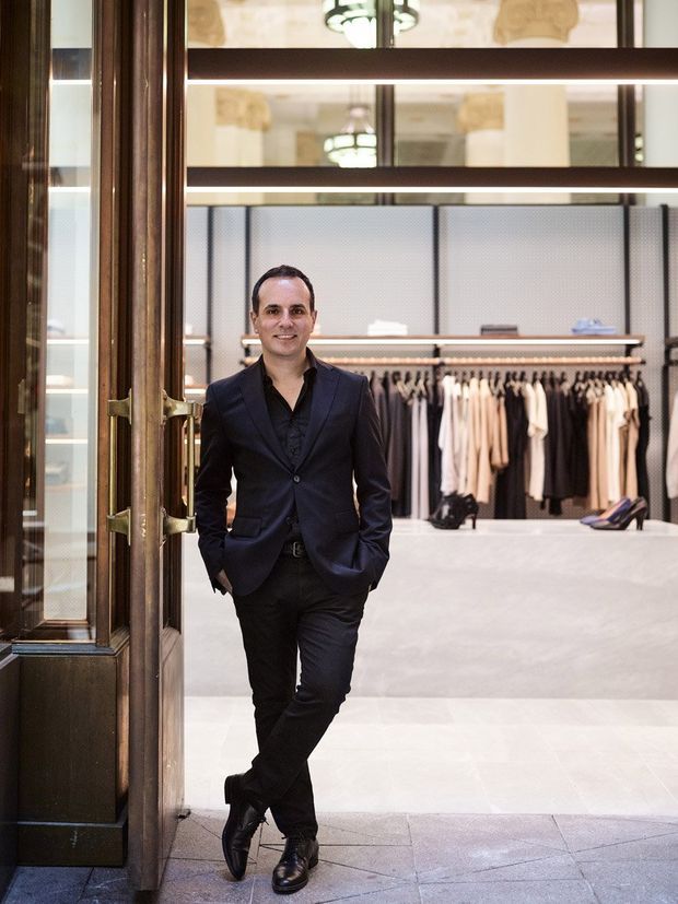 A well-dressed man stands in the open doorway of an elegant clothing boutique. The retail interior behind him features clothing racks, display shelving, shoes, pale flooring, dark timber-framed glazing and doors, and decorative pendant lighting. The image is a portrait-oriented commercial interior photograph.