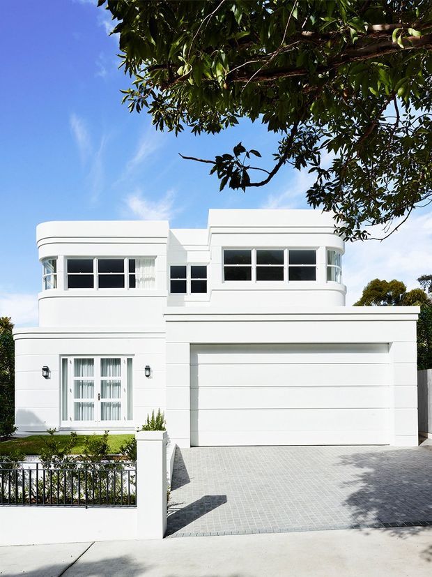 Street-facing photograph of a refined white two-storey residence with rounded corner forms, horizontal banding, large black-framed windows, a broad integrated garage, and a paved driveway. A low front fence, small landscaped garden, mature tree canopy, and clear blue sky frame the facade.