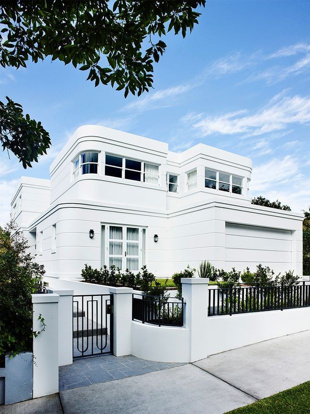 Street-facing view of a refined white two-storey residence with rounded corners, horizontal wall banding, curved upper-level windows, a concealed garage, and a formal front garden behind black metal fencing. The facade is photographed in bright daylight against a blue sky.