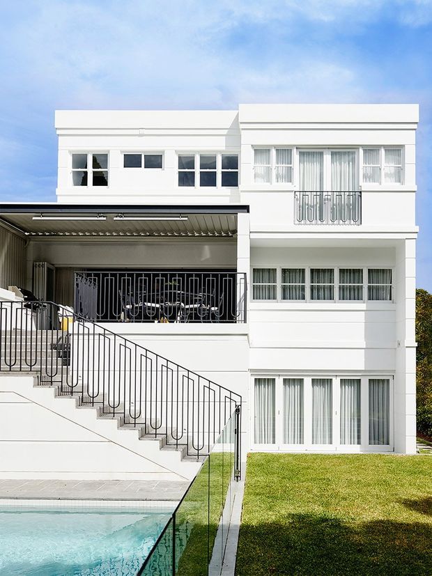 Exterior view of a large white multi-storey residence with a flat roof and strong horizontal banding. The façade features extensive white-framed glazing, sheer curtains, black metal balcony and stair balustrades, a covered outdoor entertaining area, a manicured lawn, and the edge of a swimming pool in the foreground. Bright contemporary urban residential architecture.