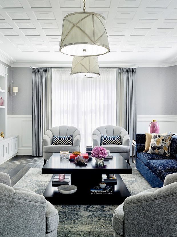 Bright, refined living room with a symmetrical seating arrangement, two grey armchairs facing a large black rectangular coffee table, a deep blue sofa, and layered patterned cushions. The room features grey paneled walls with white wainscoting and trim, full-height sheer and striped curtains, a decorative coffered ceiling, and two large drum pendant lights. A monochromatic grey rug and black furniture are accented by pink flowers and accessories.