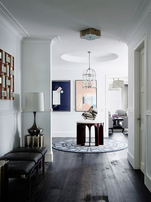 A refined residential hallway or entry passage with white paneled walls, decorative cornices and architraves, dark timber flooring, and a black leather bench. A built-in open display shelf, sculptural table lamp, framed artwork, pendant lights, and a round geometric rug add an art-deco-inspired character. The hallway opens toward a furnished living area visible in the background.