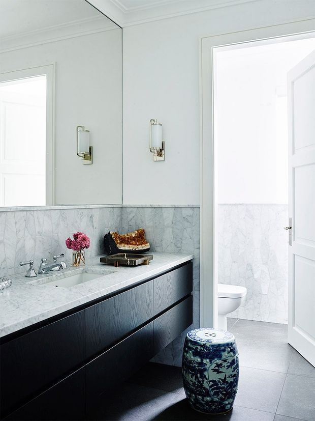 Bright, refined bathroom with a large wall-to-wall mirror, white-painted walls and trim, marble vanity countertop and matching half-height marble wall finish. The dark timber vanity has integrated drawers, chrome tapware, and wall sconces. A separate toilet is visible through the open doorway, with a decorative blue ceramic stool in the foreground.